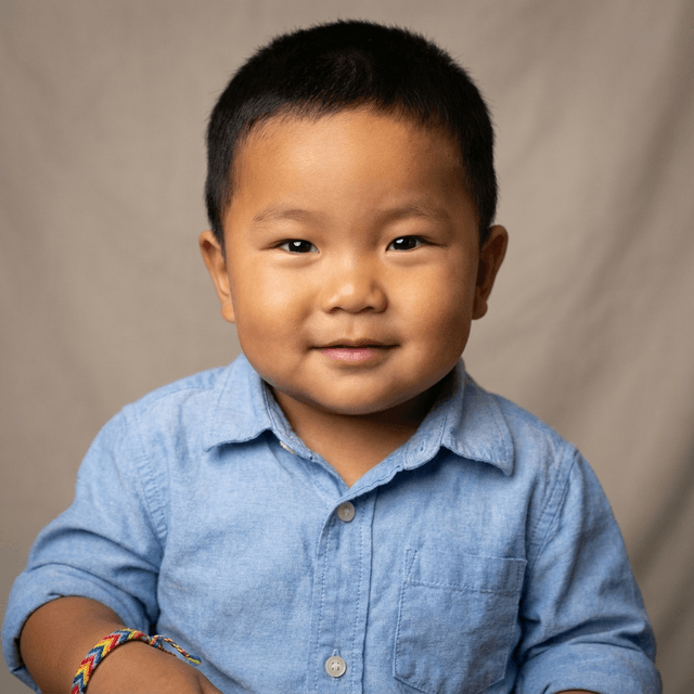 A young boy with short dark hair wearing a light blue button-up shirt and a colorful woven friendship bracelet, smiling gently.