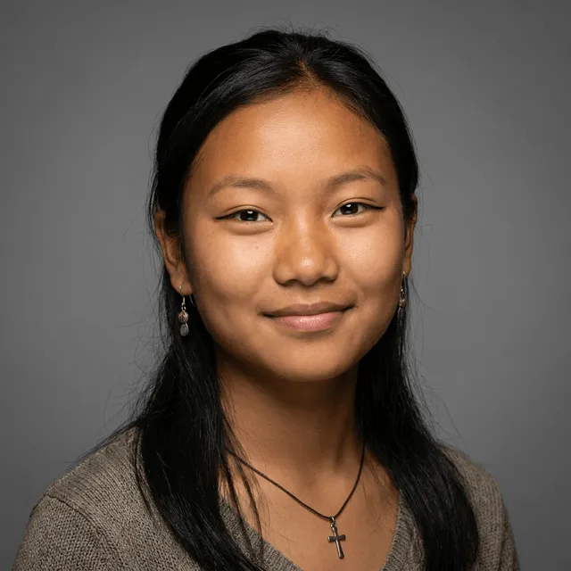 A young woman with long black hair, dangling earrings, and a small cross pendant necklace, wearing a knit top and smiling warmly at the camera.