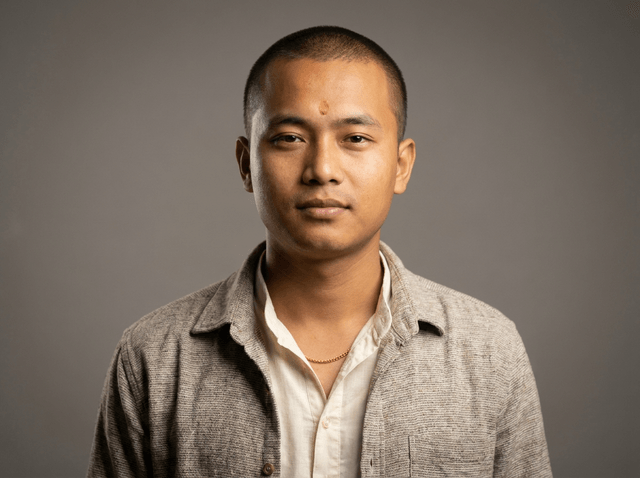 A young man with very short buzzed hair, a small reddish mark on his forehead, and a thin gold chain necklace, wearing a cream shirt under a textured grey jacket, with a neutral, calm expression.
